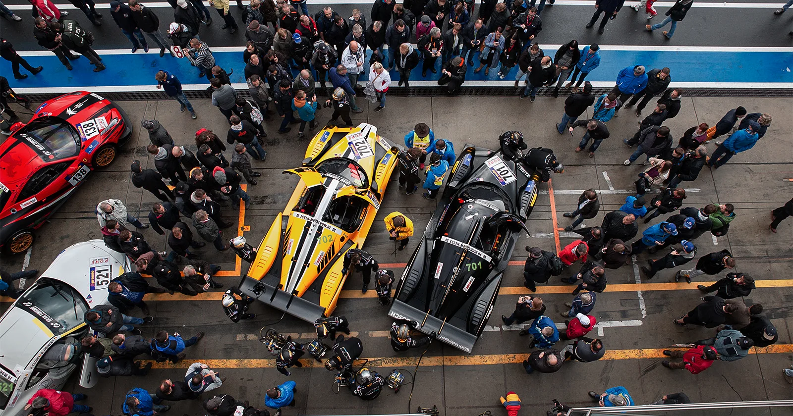 Top view of two race cars on a track with drivers and several people standing trackside watching them